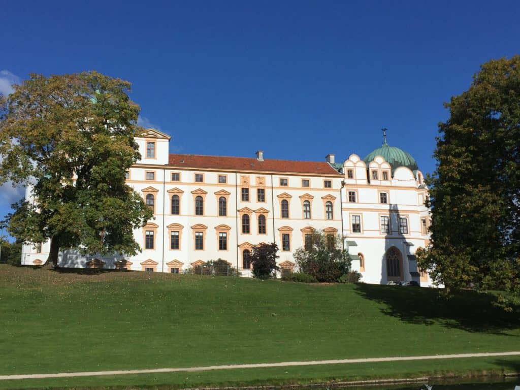 A picture of Celle castle, a place that is easily reachable even if you only have 2 days in Hannover.  It's white with orange borders around the windows and an orange roof.  It has a small green dome.  The building sits on a grassy incline with trees to the side.