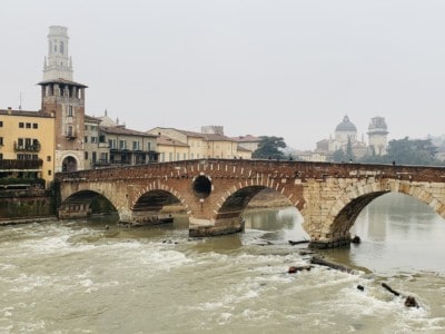 A picture of the Ponte Pietra bridge.  This has brown and yellow bricks and is arched  You can see houses in the background and a tower, as well as the duomo in the distance