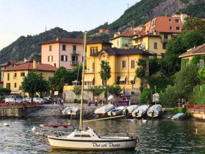 The port of Varenna, a small port with boats on the shoreline, pretty colourful houses and an outcrop with bushes behind. There is a boat bobbing on the water on the foreground.