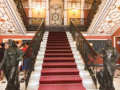 A staircase inside Achilleoin Palace.  It has a red carpet and ornate patterned ceilings above.  There is a statute flanking each side of the base.