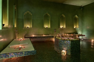 The second room in the Hammam De La Rose.  You can see a dimly lit room with cut out arches in the walls.  There are beaches along the side of the wall and a font in the middle with a lamp at the base of it.