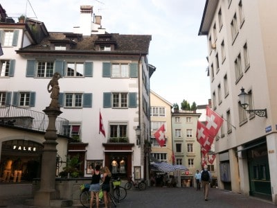 A street in the old town. There are building with the Swiss flag flying from them and a fountain in the street on the left.